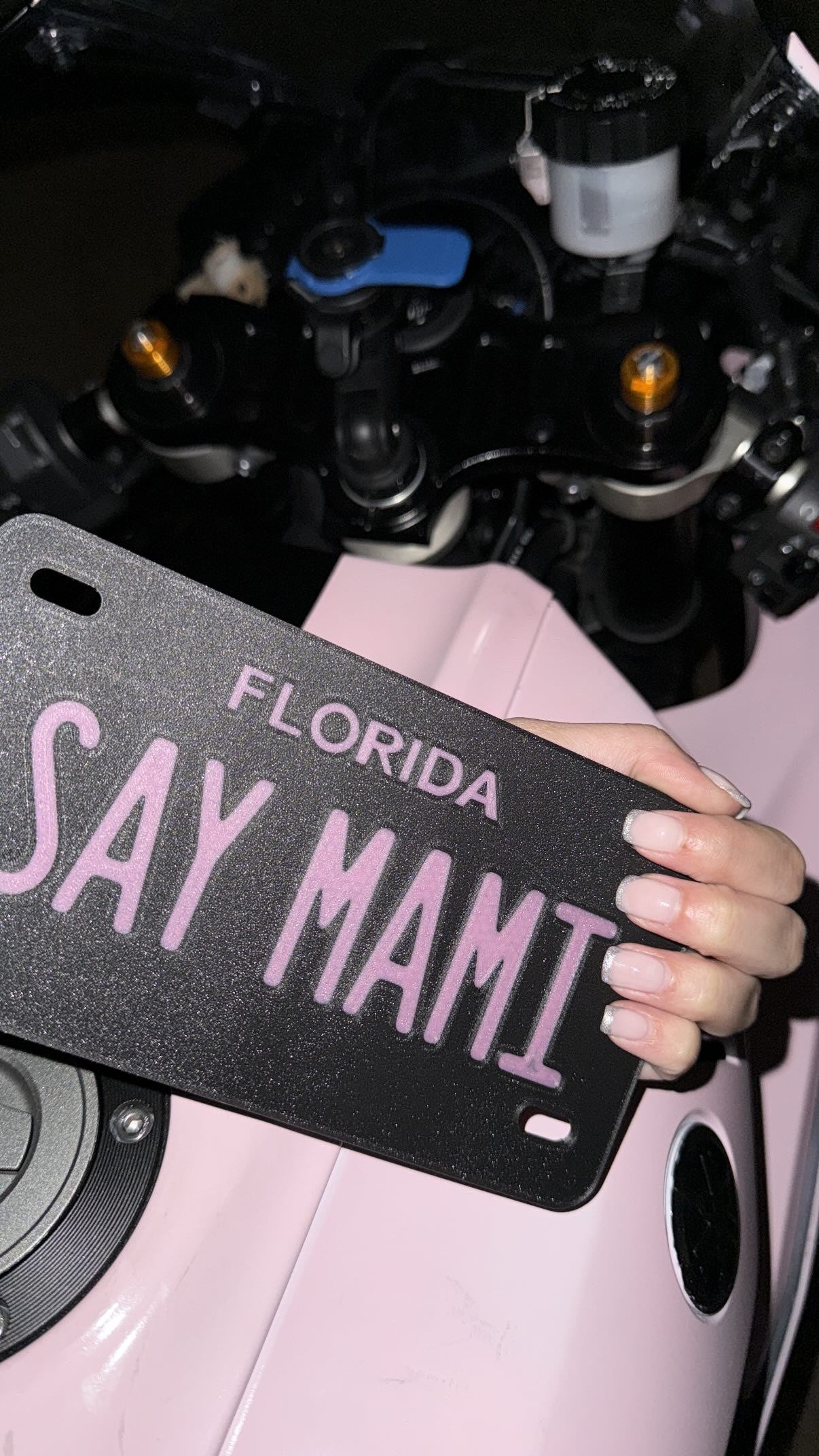 Custom motorcycle vanity plate, pink and black with text "SAY MAMI"